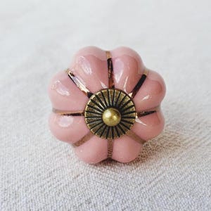May include: A pink ceramic cabinet knob in a flower shape. The knob has a bronze centre and bronze accents. The knob is set against a white background.