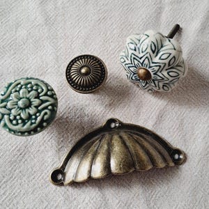 May include: A selection of cabinet hardware, including ceramic and metal knobs and a pull. The knobs display floral and geometric patterns in green, white, and bronze. The pull has a fan-shaped design.
