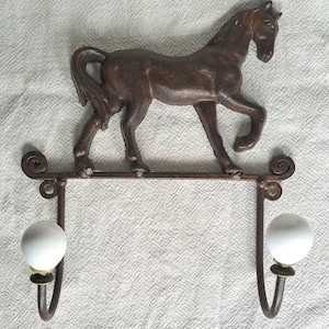 May include: A rustic wall hook with a brown horse figure. The horse is mounted on a dark metal bar with two curved hooks and white ceramic ball details. The design is equestrian-themed, with a vintage feel.