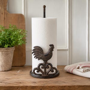 May include: A vertical paper towel holder made of dark brown cast iron featuring a rooster figure standing on a decorative scrollwork base. The holder has a central rod that supports a roll of white paper towels with a subtle embossed pattern.