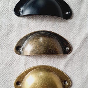May include: Three metal cup pull handles in black, bronze, and brass. Each handle has a semi-circular design with two screw holes for mounting. The handles are arranged vertically against a neutral fabric background.