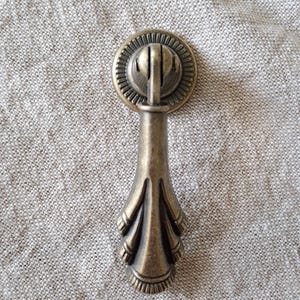 May include: Antique brass cabinet pull with a decorative, ornate design. The pull has a round top with a raised centre and a long, slender handle with a decorative, floral-like design at the bottom.