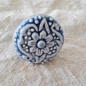 May include: A round, blue and white ceramic knob with a floral design. The knob features a raised, detailed pattern of a flower and swirling accents. The knob is likely for a cabinet or drawer.