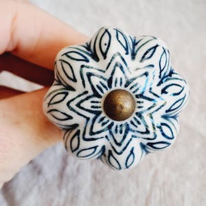 May include: A white ceramic cabinet knob with a navy blue floral pattern. The knob features a brass-coloured centre and a rounded shape. The symmetrical floral design adds a decorative element to furniture. The knob is approximately 4 cm in diameter.