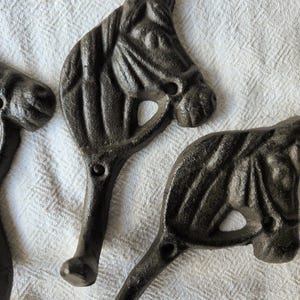 May include: A set of three dark brown cast iron wall hooks, each featuring a detailed relief of a horse head with a mane and bridle. The hooks extend downwards from the base of the horse head design, providing a functional element for hanging items.