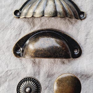 May include: A selection of antique bronze cabinet hardware, including two cup pulls and two knobs. The cup pulls have a shell-like design, while the knobs feature a sunburst and a smooth, rounded surface.