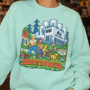 Puede incluir: Una sudadera de cuello redondo en color verde menta con un estampado gráfico colorido que muestra un barrio residencial junto a un centro de datos de IA. La ilustración incluye personajes de dibujos animados, un perro y el texto "AI DATA CENTER" y "RESPECT THE LOCALS."