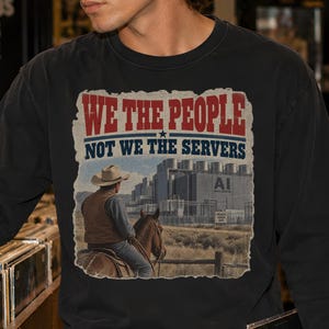 May include: A black long-sleeve shirt featuring a graphic print of a cowboy on horseback looking toward a large industrial building. The text reads WE THE PEOPLE NOT WE THE SERVERS. The graphic includes a red and blue color scheme.