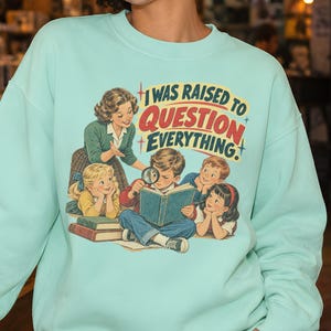 May include: A light mint green crewneck sweatshirt featuring a vintage-style graphic of a teacher and four children reading books. The text "I WAS RAISED TO QUESTION EVERYTHING" is printed in red and blue above the illustration of the group.