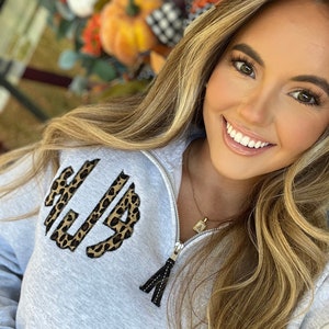 May include: A gray sweatshirt with a leopard print monogram on the front. The sweatshirt has a zipper and a black tassel pull. The model is wearing a gold necklace with a small pendant.
