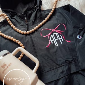 Puede incluir: Chaqueta negra con capucha con un lazo rosa y monograma con las letras "APK". También se ve un collar de cuentas de madera y una taza de viaje beige Stanley con el texto "ivory lee".