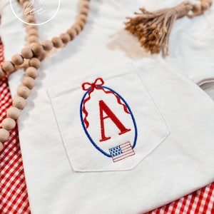 Op de afbeelding: Witte T-shirt met geborduurde zak, een rood-wit-blauwe Amerikaanse vlag en de letter A in een rode ovaal met een blauwe omtrek. Het T-shirt ligt op een rood-wit geruite achtergrond.