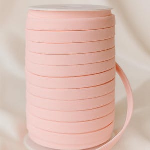 May include: A spool of light pink bias tape or ribbon wound tightly around a white plastic core. The word PINK is printed in bold black capital letters above the spool. The material has a smooth, matte texture and a consistent width.
