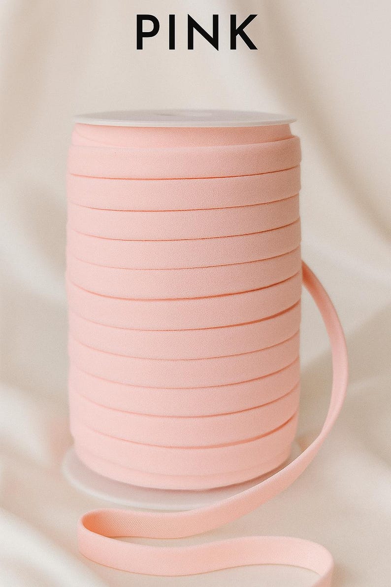 May include: A spool of light pink bias tape or ribbon wound tightly around a white plastic core. The word PINK is printed in bold black capital letters above the spool. The material has a smooth, matte texture and a consistent width.