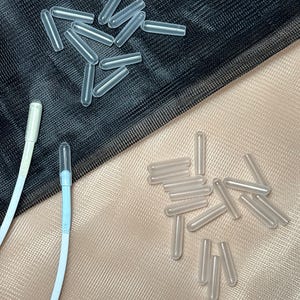 May include: Two piles of clear plastic caps, one pile on a black mesh surface and the other on a beige mesh surface. The caps are cylindrical and translucent.