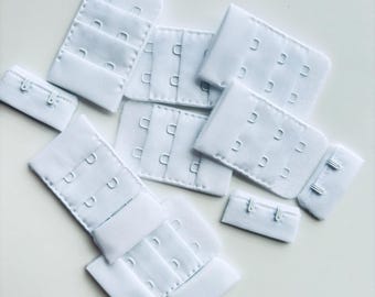 May include: A collection of white bra hook and eye closures in various sizes. The closures are made of a soft, textured fabric and feature multiple hook and eye sets for adjustable fit. Ideal for lingerie and sewing projects.