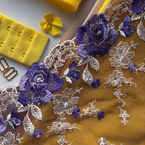 May include: Close-up of various sewing supplies, including yellow elastic, a hook and eye closure, and a small yellow bow. The image also features a piece of fabric with intricate purple floral embroidery and delicate white lace detailing.