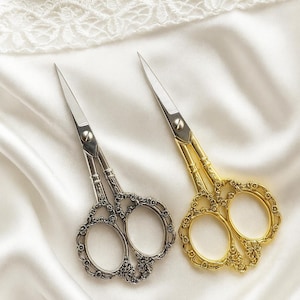 Op de afbeelding: Twee decoratieve scharen met sierlijke handvatten. Eén paar heeft een zilveren afwerking, de andere is goudkleurig. De bladen zijn zilver en puntig. De scharen worden gepresenteerd op een witte, zijdeachtige stof.