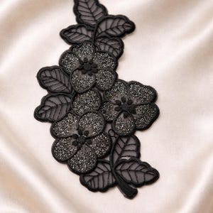 May include: Black floral applique with glitter accents. The applique features detailed leaves and flowers, perfect for embellishing garments or crafts. The design is set against a cream-colored satin background.