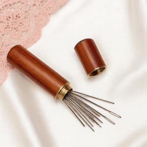 May include: A wooden needle case with a brass-colored rim, open to reveal a bundle of needles. The case is cylindrical and made of a dark wood. The lid is separate. The background is a white, silky fabric.