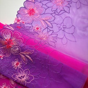 Op de afbeelding: Paarse en roze ombre bloemenkant met een gegolfde rand. De kant is geborduurd met delicate bloemen en bladeren.