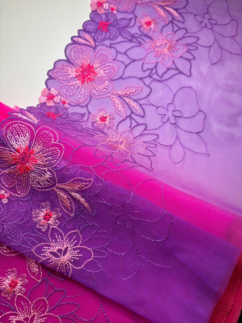 Op de afbeelding: Paarse en roze ombre bloemenkant met een gegolfde rand. De kant is geborduurd met delicate bloemen en bladeren.
