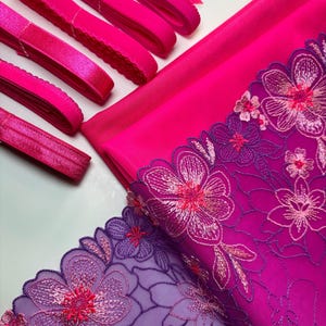 Op de afbeelding: Een set roze stof en kant voor knutselprojecten. De set bevat een stuk roze stof, een stuk paarse kant met roze bloemenborduurwerk en verschillende roze elastiekjes.