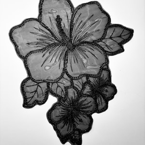 May include: Black and white floral applique featuring a large hibiscus flower with detailed black stitching and beaded edges. The design includes multiple flower blossoms and leaves, suitable for fabric embellishment or crafting projects.