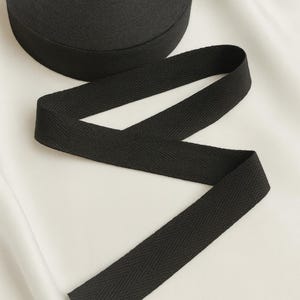 May include: A roll of black herringbone twill tape with a length of the tape unrolled. The tape is a solid black color and has a textured surface. The tape is likely used for sewing, crafting, or other textile projects. The brand name "Charmlace Atelier" is visible.