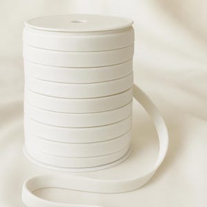 May include: A spool of white elastic ribbon, with the word "WHITE" in gray text at the top. The ribbon is neatly coiled, with a length of the ribbon extending outwards. The background is a soft, white fabric.