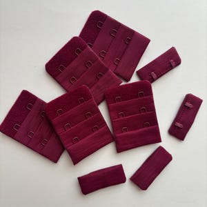 May include: Burgundy bra hook and eye closures in various sizes. The closures are made of fabric and feature metal hooks and eyes. The image shows several sets of closures, including hook and eye strips and smaller hook and eye pieces.