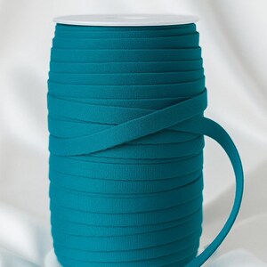 May include: A spool of teal flat elastic cord wound tightly around a white plastic cylinder. The cord has a matte, textured surface and is wrapped in multiple horizontal layers, with one end extending outward from the main spool.
