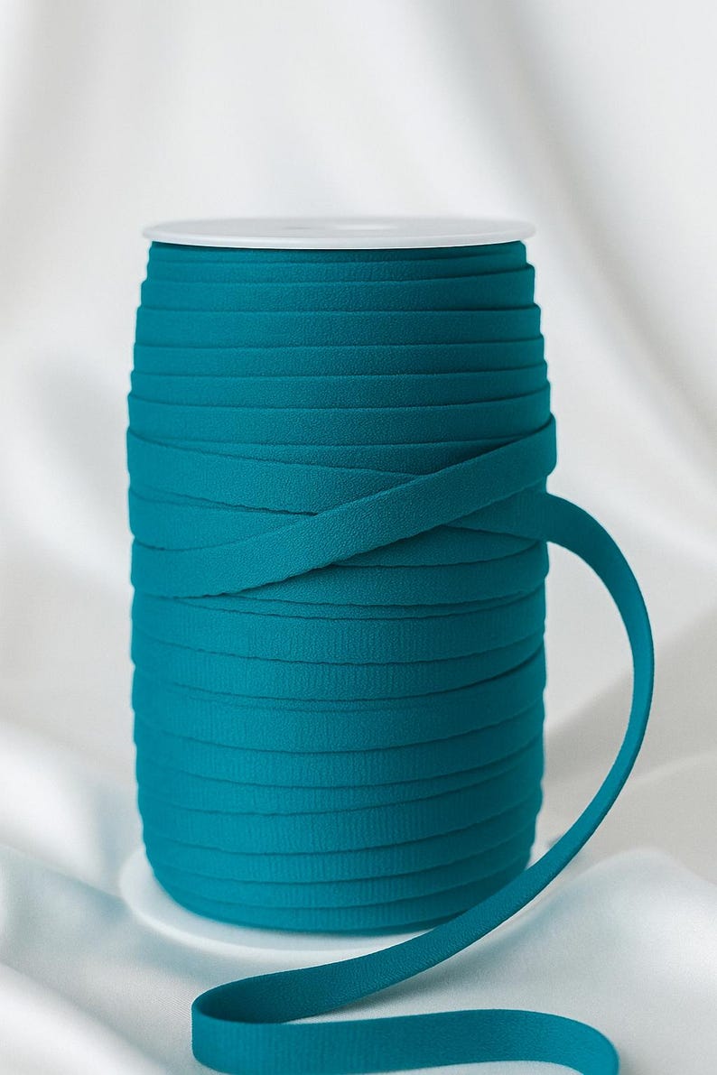 May include: A spool of teal flat elastic cord wound tightly around a white plastic cylinder. The cord has a matte, textured surface and is wrapped in multiple horizontal layers, with one end extending outward from the main spool.