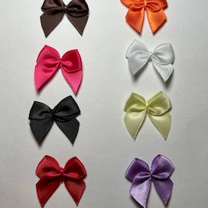 Puede incluir: Surtido de diez pequeños lazos de cinta de satén en varios colores, incluyendo marrón, naranja, blanco, negro, amarillo, rojo y morado.