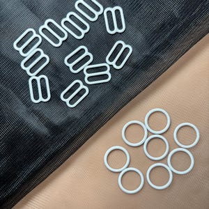 May include: White plastic bra strap sliders and rings. The sliders are rectangular and the rings are round. They are on a black and beige fabric background.