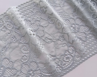 May include: A wide strip of light gray lace fabric featuring a detailed floral pattern with intricate mesh sections and scalloped edges. The delicate textile displays a repeating botanical design with openwork elements throughout the entire length.
