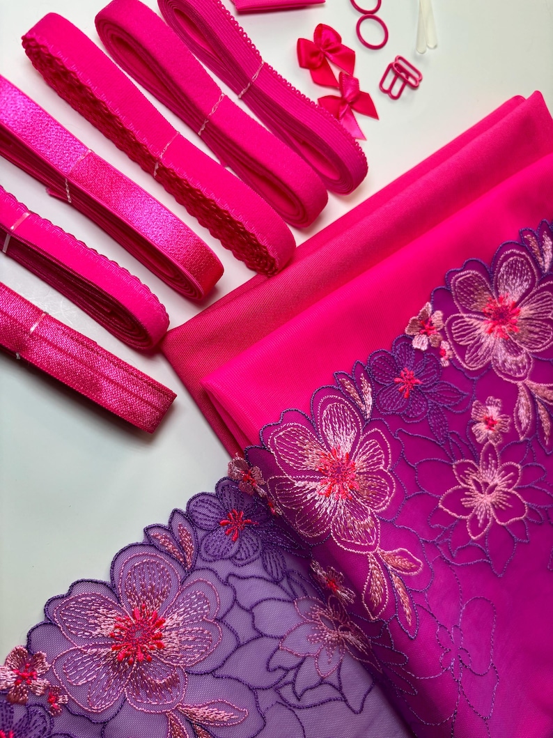 Op de afbeelding: Een set met roze elastische trim, roze stof en paars-roze bloemenkant. De stof is effen roze en de kant heeft een verloop van paars naar roze.