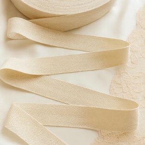 May include: A roll of cream-colored herringbone ribbon with a length of ribbon unraveled. The ribbon is made of a woven fabric and is displayed on a white surface. The text "Charm Lace Atelier" is visible.