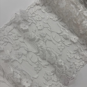 May include: Close-up of a white floral lace fabric. The lace features a delicate pattern of flowers and leaves with silver metallic thread accents. The fabric is soft and has a scalloped edge, suitable for various craft projects.
