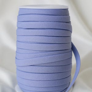 May include: A spool of light periwinkle blue bias tape. The tape is tightly wound on a white spool, with a length of tape extending from the side. The fabric has a soft, textured appearance, suitable for sewing and crafting projects.