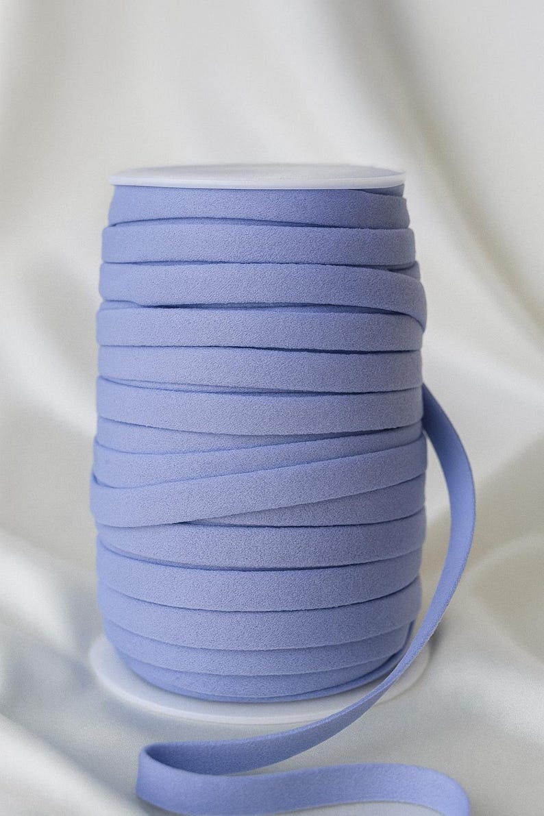 May include: A spool of light periwinkle blue bias tape. The tape is tightly wound on a white spool, with a length of tape extending from the side. The fabric has a soft, textured appearance, suitable for sewing and crafting projects.