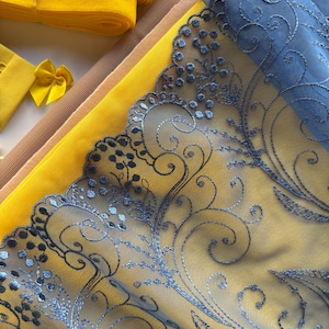 May include: A close-up of various fabrics and ribbons in shades of yellow, tan, and blue. The focal point is a piece of blue embroidered lace with a floral design. Rolls of yellow ribbon and small yellow bows are also visible.