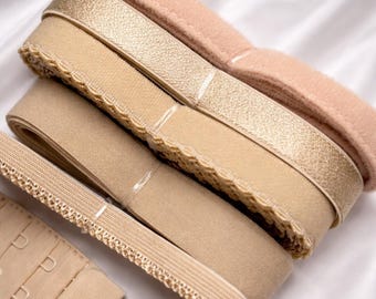 May include: An assortment of beige and gold bra-making supplies, including elastic bands, hook and eye closures, and bow accents. The materials are arranged on a white, silky surface, showcasing the various textures and shades.