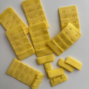 May include: A collection of yellow bra extenders, featuring multiple rows of hook-and-eye closures. The extenders are made of a soft, fabric-like material. The image shows various sizes and configurations of the extenders.