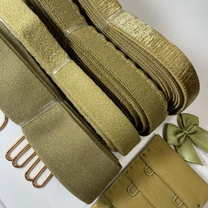 May include: An assortment of olive green bra-making supplies, including hook and eye closures, bra straps, and a small bow. The materials are neatly arranged, showcasing various textures and finishes.