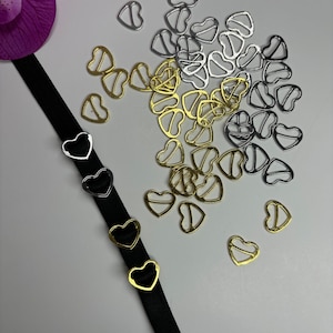 以下が含まれることがあります： 黒いリボンに3つのハート型の金属製留め具が付いているクローズアップ。留め具は金、銀、黒です。リボン周辺には、金と銀のハート型の留め具が多数散らばっています。