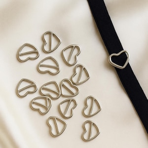 May include: Silver-toned metal heart-shaped sliders and a heart-shaped buckle on a black strap. The sliders are scattered on a white satin surface. The heart-shaped buckle is attached to the black strap.