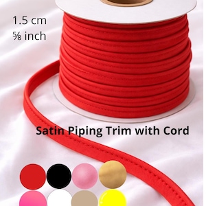 Könnte beinhalten: Eine Spule mit roter Satin-Paspel mit Kordel, 1,5 cm breit, wird gezeigt. Die Paspel ist auf einer weißen Pappspule aufgewickelt. Das Bild zeigt auch eine Farbpalette der Paspel in verschiedenen Farben, darunter Rot, Schwarz, Rosa und Gelb.