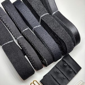 May include: An assortment of black bra-making supplies, including elastic bands, hook and eye closures, strap adjusters, and clear plastic strap sliders. The elastic bands vary in texture and width, with some having a decorative edge.