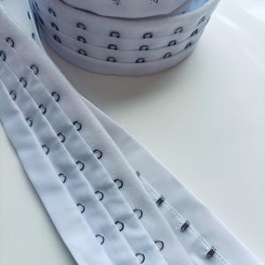 May include: White bra hook and eye closures and hook and eye tape. The closures are stacked in a pile, and the tape is laid out in rows. The metal hooks and eyes are silver.